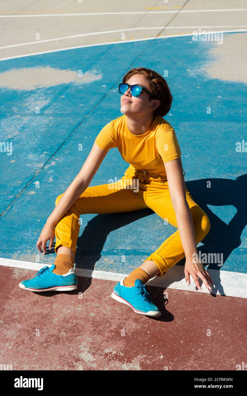 Grand angle de frowning froide femme adolescente dans des vêtements jaunes  avec des lunettes de soleil tout en étant assis seul sur un terrain de  sport bleu de basket-ball contre les bâtiments