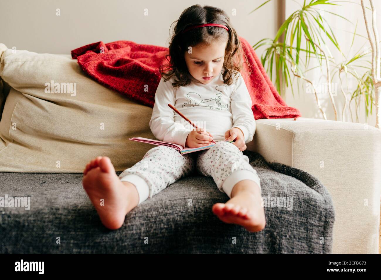 Adorable fille pieds nus dans un pyjama assis sur un canapé et dessin dans  un carnet de croquis dans un salon confortable à la maison Photo Stock -  Alamy