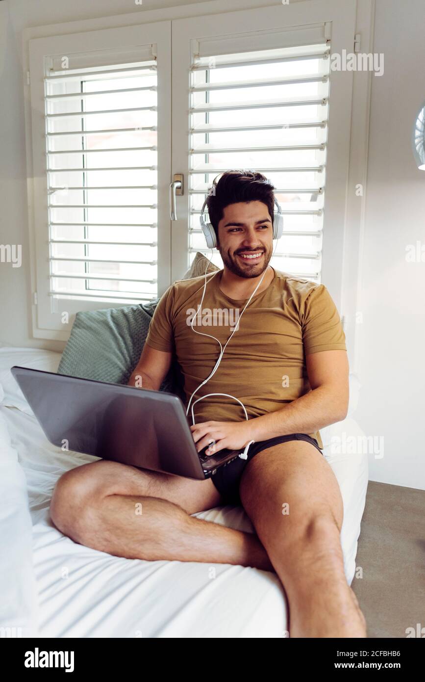 Jeune homme joyeux et réussi dans une chemise et un casque décontractés assis sur le lit avec ordinateur portable et vue sur l'extérieur Banque D'Images