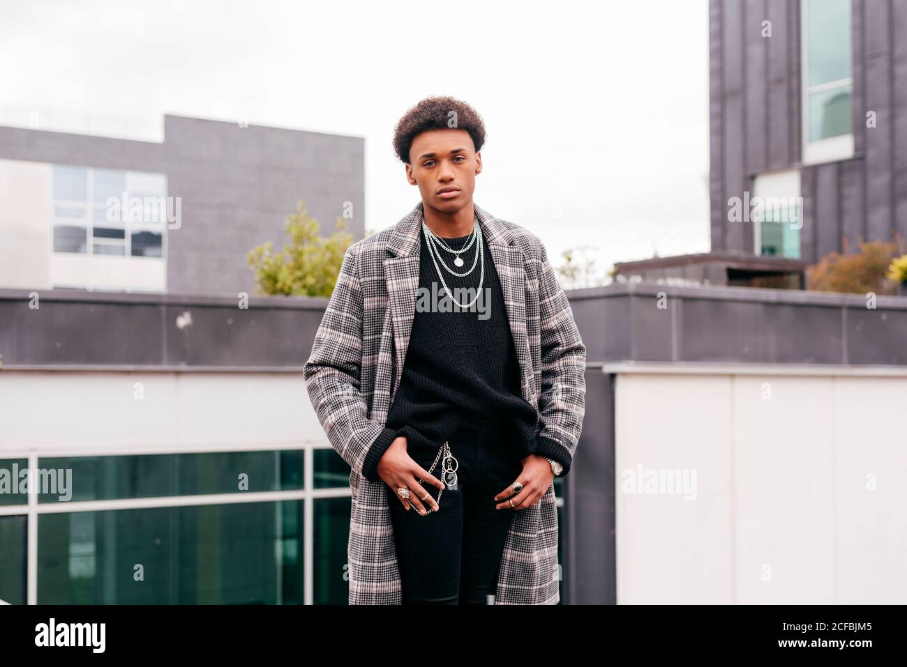 Un jeune homme à la mode afro-américain sérieux en manteau à carreaux  tendance et un pantalon serré qui regarde l'appareil photo tout en se  tenant contre un vêtement moderne bâtiments de la