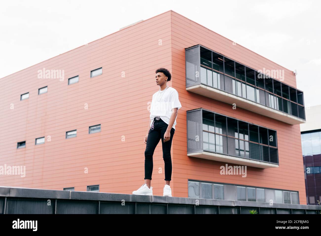 Vue d'en dessous de côté de jeune homme afro-américain élégant une chemise blanche et un pantalon serré blanc baskets debout sur une clôture contre un bâtiment rose de la ville Banque D'Images