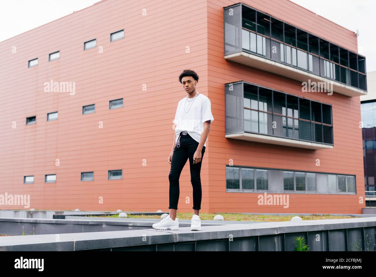 Vue d'en dessous de côté de jeune homme afro-américain élégant regarder l'appareil photo dans une chemise blanche et un pantalon serré avec baskets blanches sur la clôture contre le bâtiment rose de la ville Banque D'Images