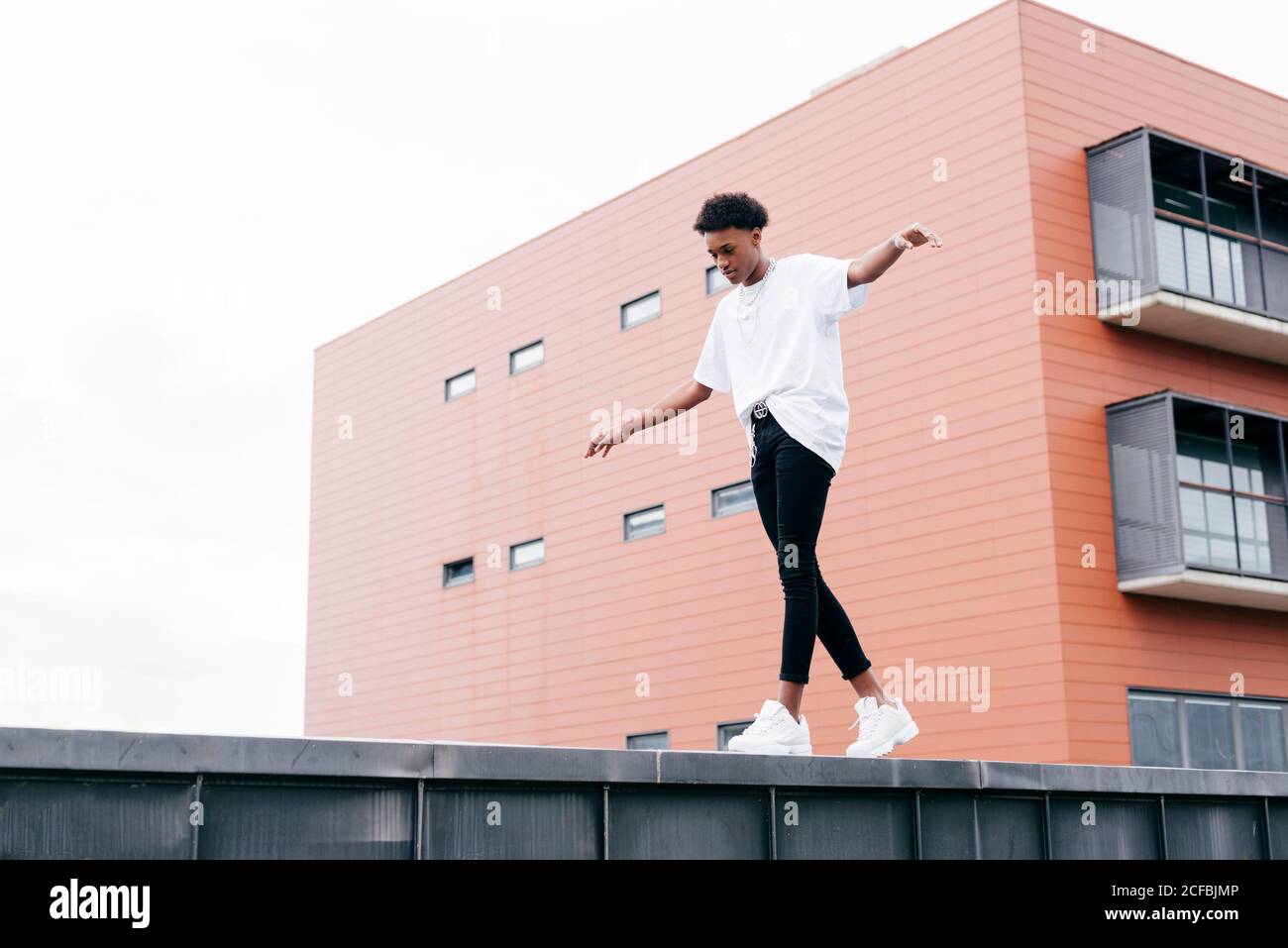 Vue d'en dessous de côté de jeune homme afro-américain élégant une chemise blanche et un pantalon serré blanc baskets debout sur une clôture contre un bâtiment rose de la ville Banque D'Images