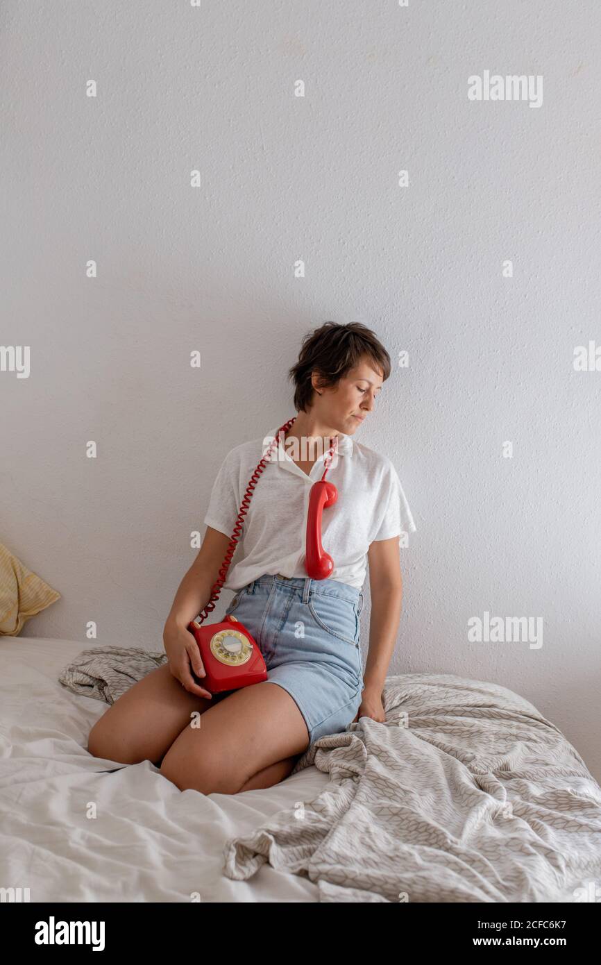 Femme indifférente avec le combiné du téléphone rouge près de l'oreille écouter pour dialoguer en partenariat tout en étant assis sur le lit contre le mur blanc dans la chambre Banque D'Images