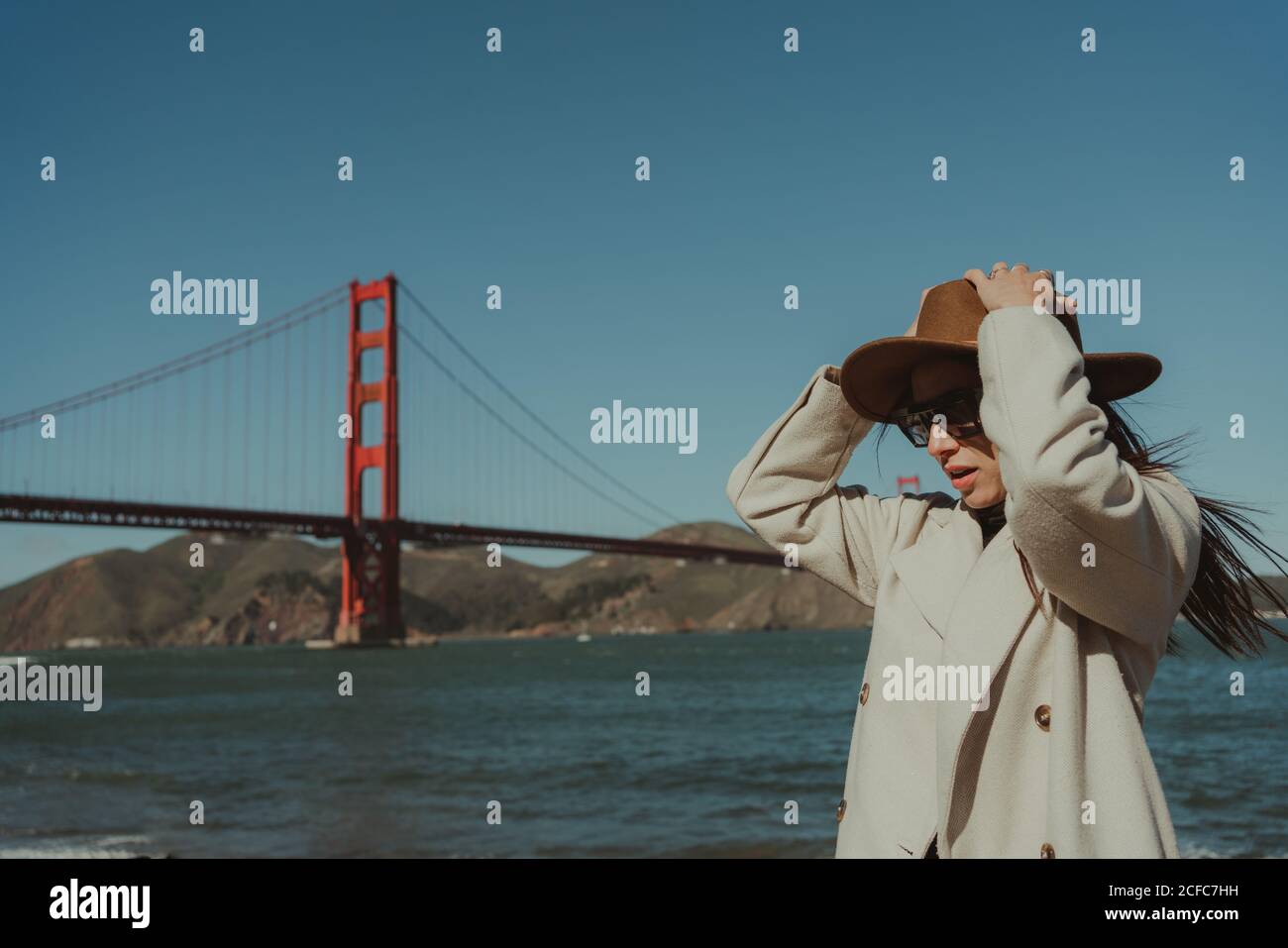 Vue latérale d'une jeune femme avec chapeau et tenue tendance Et des lunettes de soleil se tenant sur le remblai contre le pont du Golden Gate dedans Californie en journée ensoleillée Banque D'Images