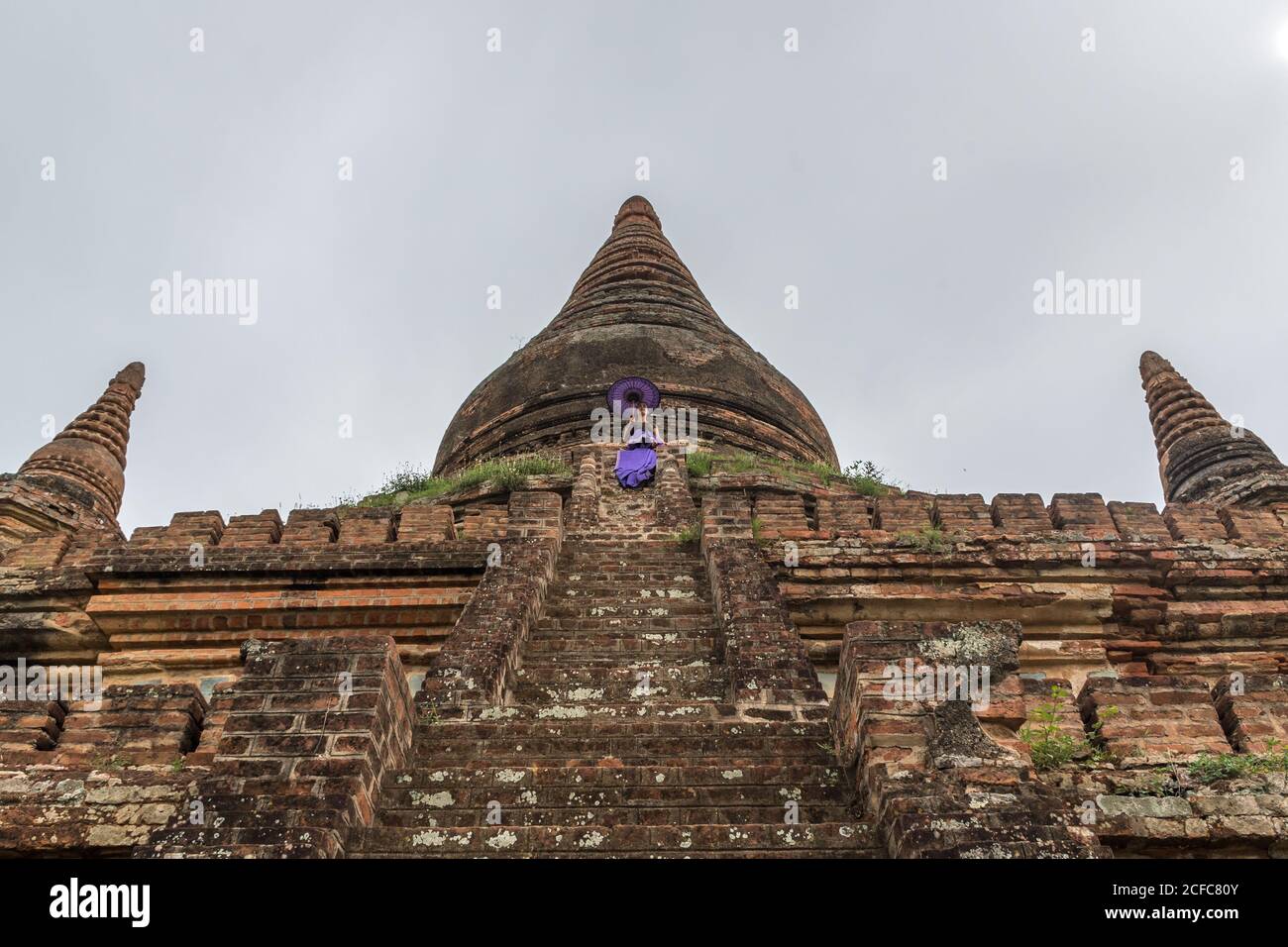 De dessous de l'ancien temple birman en pierre à Bagan contre ciel gris nuageux avec robe violette pour femme tenant le violet parasol sur les escaliers Banque D'Images