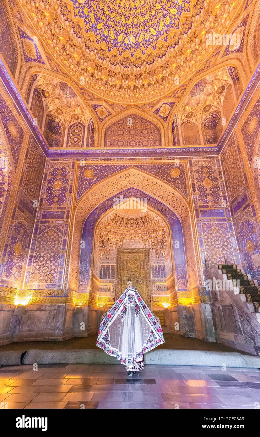 Vue arrière de la femme anonyme en robe traditionnelle se tenant au milieu d'un bâtiment ornemental illuminé lors d'une visite du Registan à Samarkand, Ouzbékistan Banque D'Images