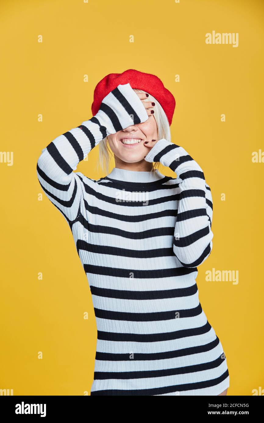 Blonde gaie femme en chemisier rayé et casquette française rouge souriant avec les mains sur le visage sur fond jaune Banque D'Images
