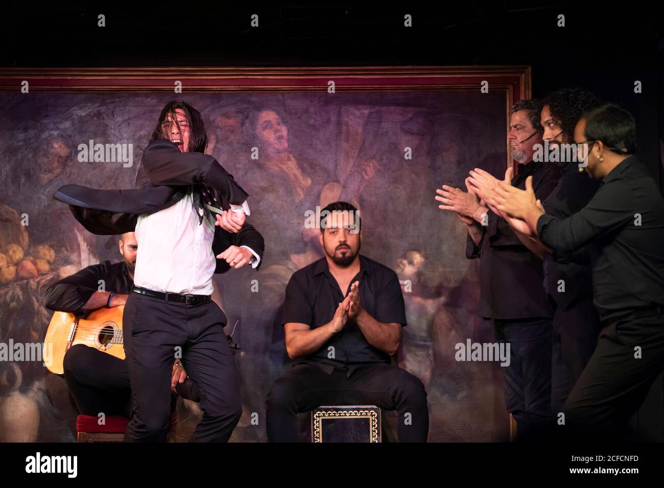 Homme en costume noir dansant flamenco près de musiciens hispaniques masculins pendant la performance contre la peinture sur scène sombre Banque D'Images