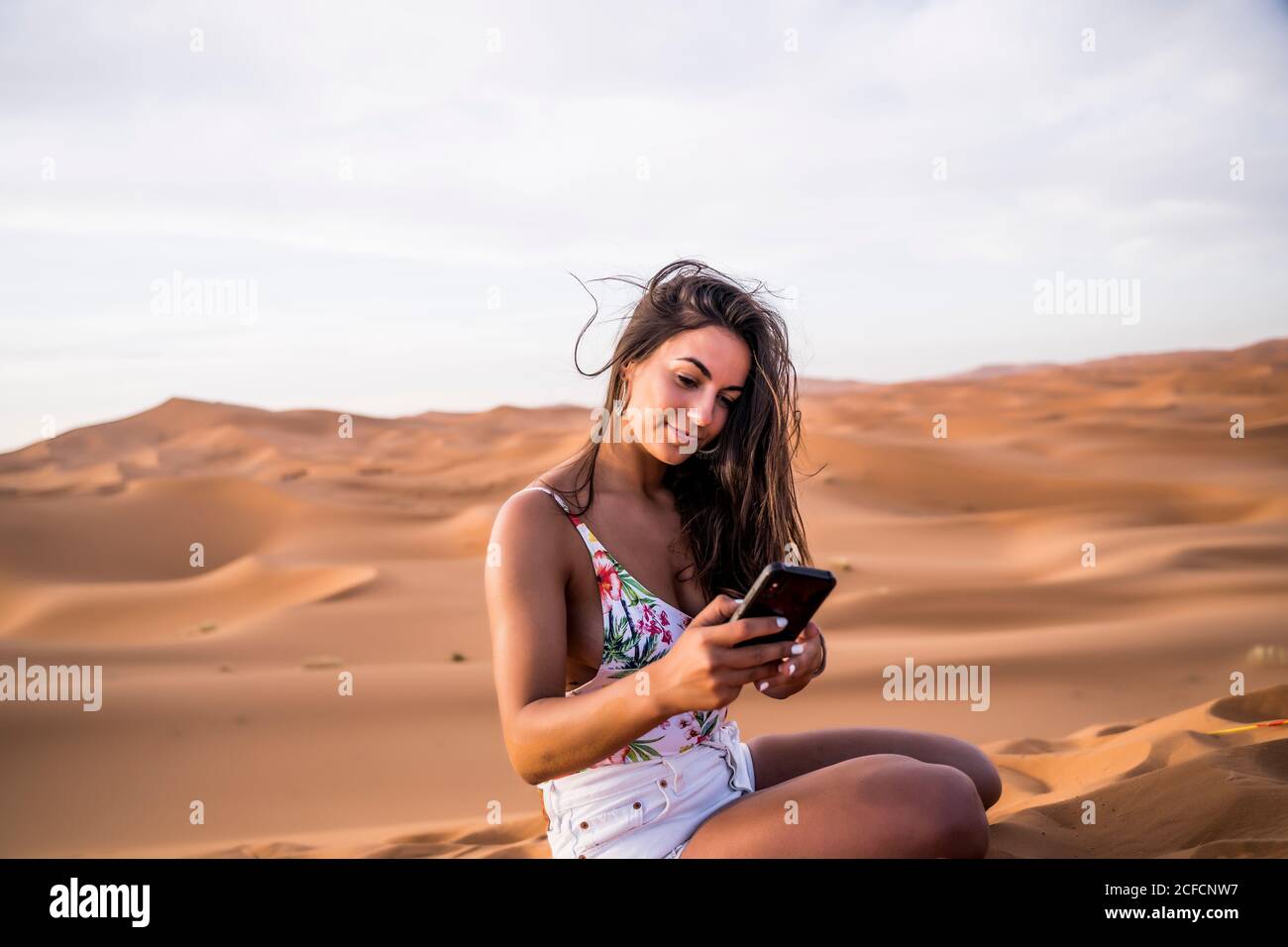 Jeune femme prenant le téléphone tout en étant assise au milieu du désert sablonneux, au Maroc Banque D'Images