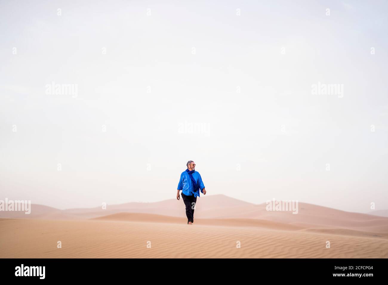 Homme mûr et élégant à barbe, regardant loin en marchant dans le sable Désert contre ciel gris au Maroc Banque D'Images