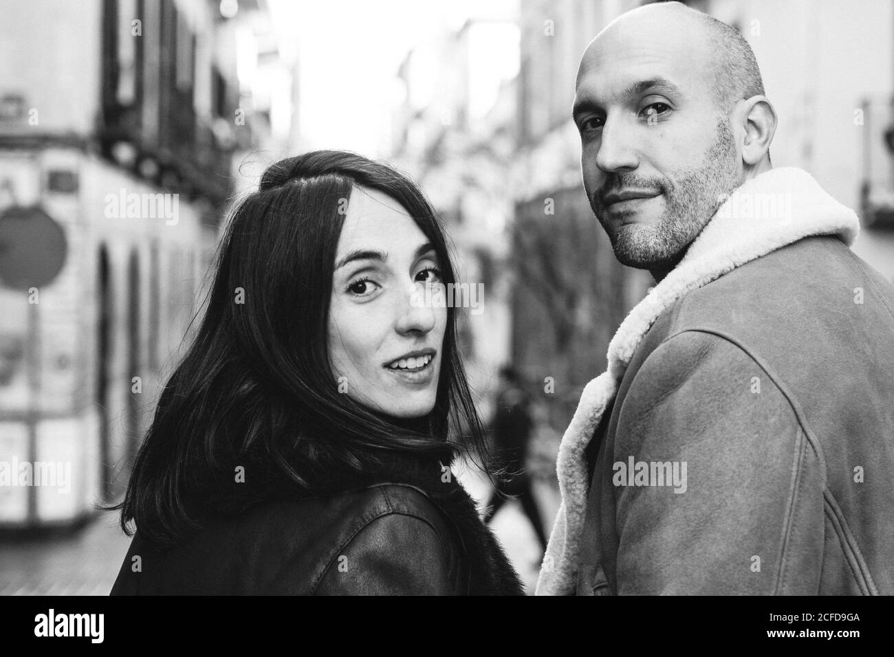 Vue arrière en noir et blanc d'un homme d'adulte chauve et d'une belle femme souriant et regardant l'appareil photo sur l'épaule tout en marchant sur une rue floue de la ville pendant la date Banque D'Images