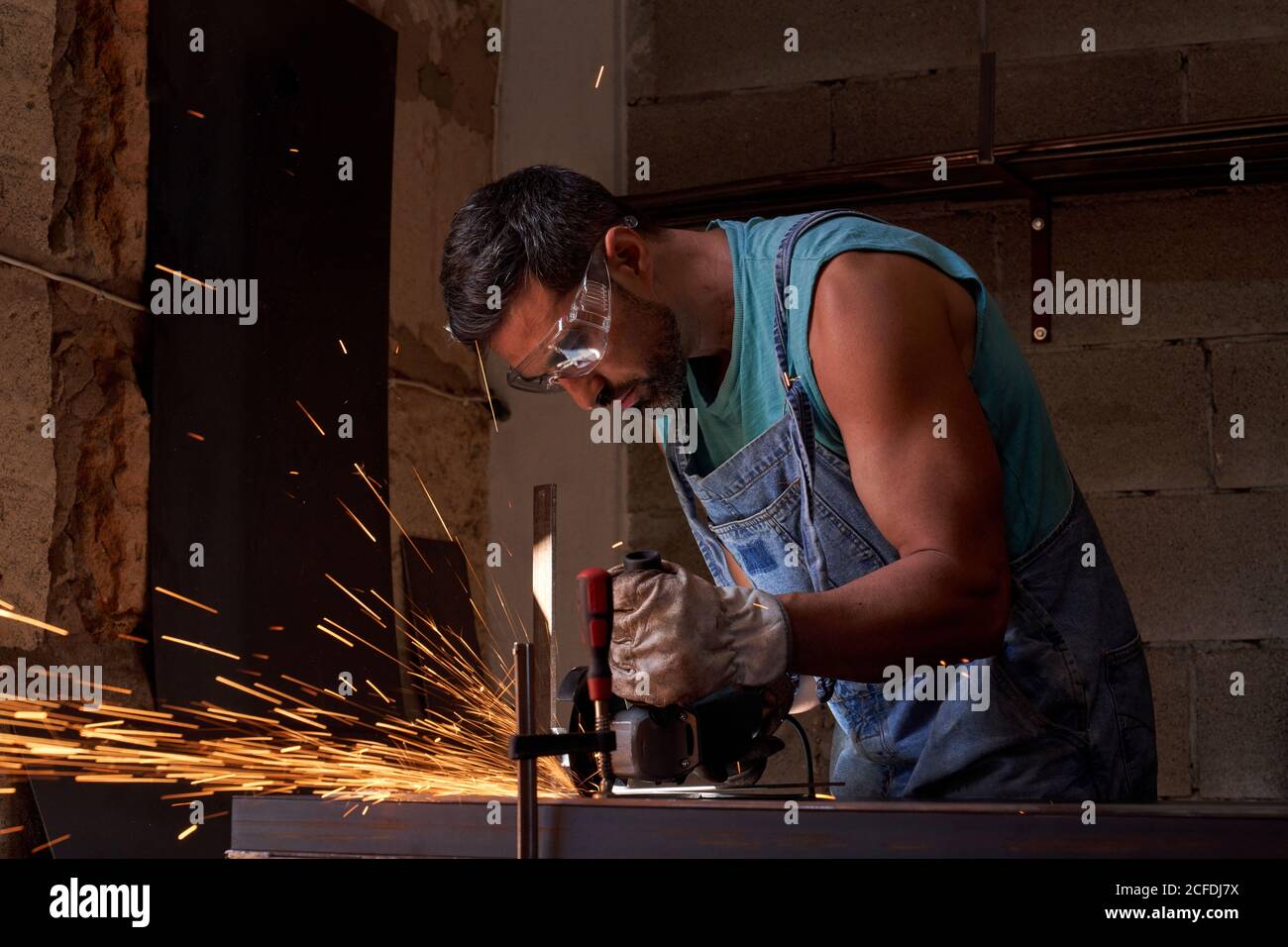 Vue latérale du travailleur dans les lunettes de protection et les gants de coupe métal avec moulin à étincelles volantes pendant les travaux en atelier Banque D'Images