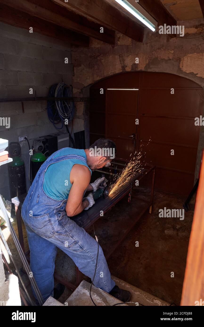 Vue latérale du travailleur dans les lunettes de protection et les gants de coupe métal avec moulin à étincelles volantes pendant les travaux en atelier Banque D'Images