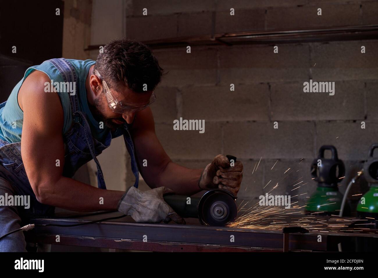 Vue latérale du travailleur dans les lunettes de protection et les gants de coupe métal avec moulin à étincelles volantes pendant les travaux en atelier Banque D'Images