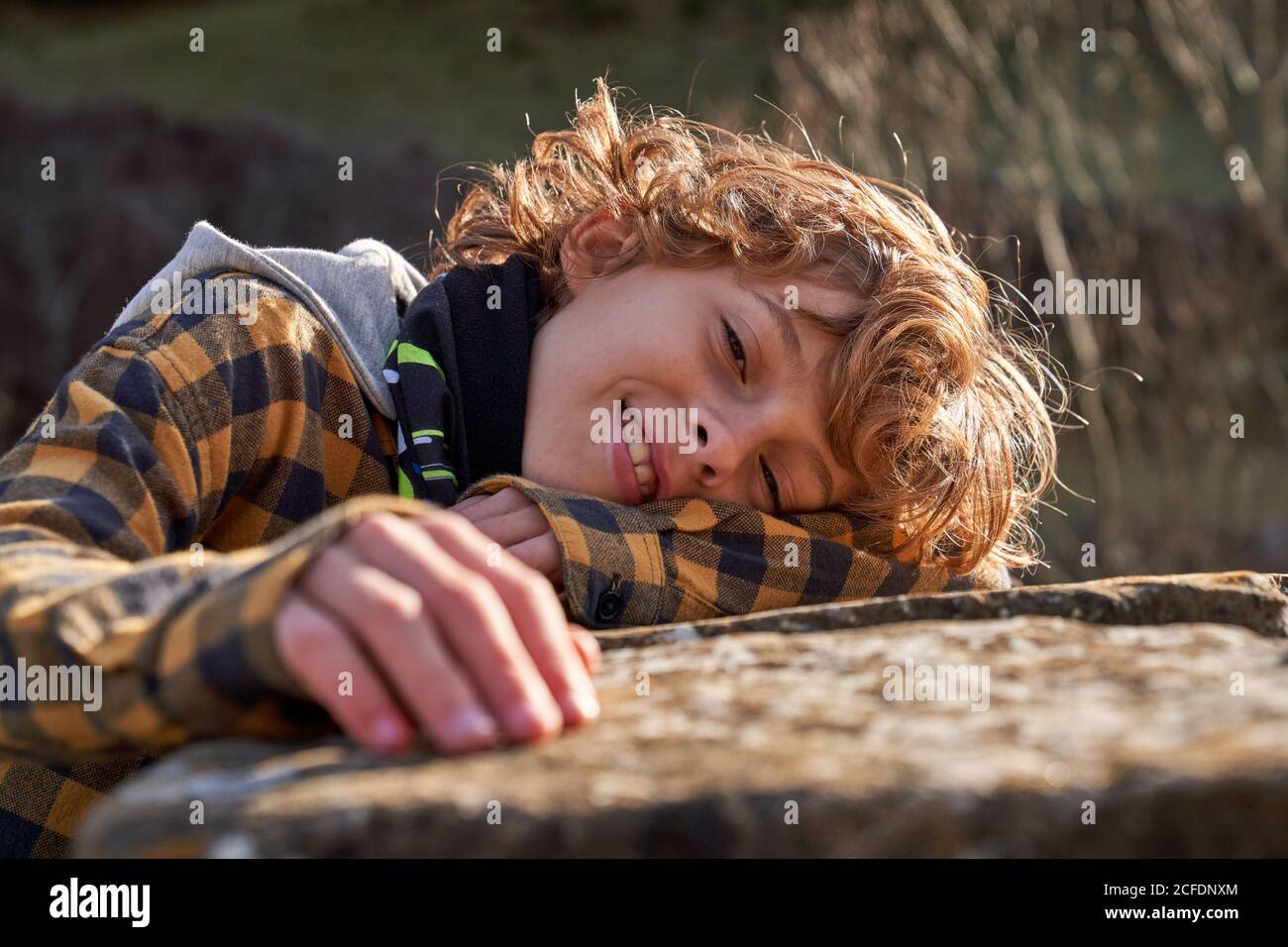 Vue latérale de l'enfant tendre curly pensif couché main sur la clôture en pierre dans la vallée de la forêt en regardant la caméra Banque D'Images