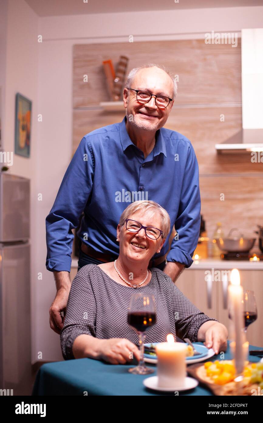 Femme handicapée excitée en fauteuil roulant et son mari derrière elle regardant la caméra. Joyeux couple senior joyeux dînant ensemble dans la cuisine confortable, en appréciant le repas, célébrant leur anniversaire. Banque D'Images