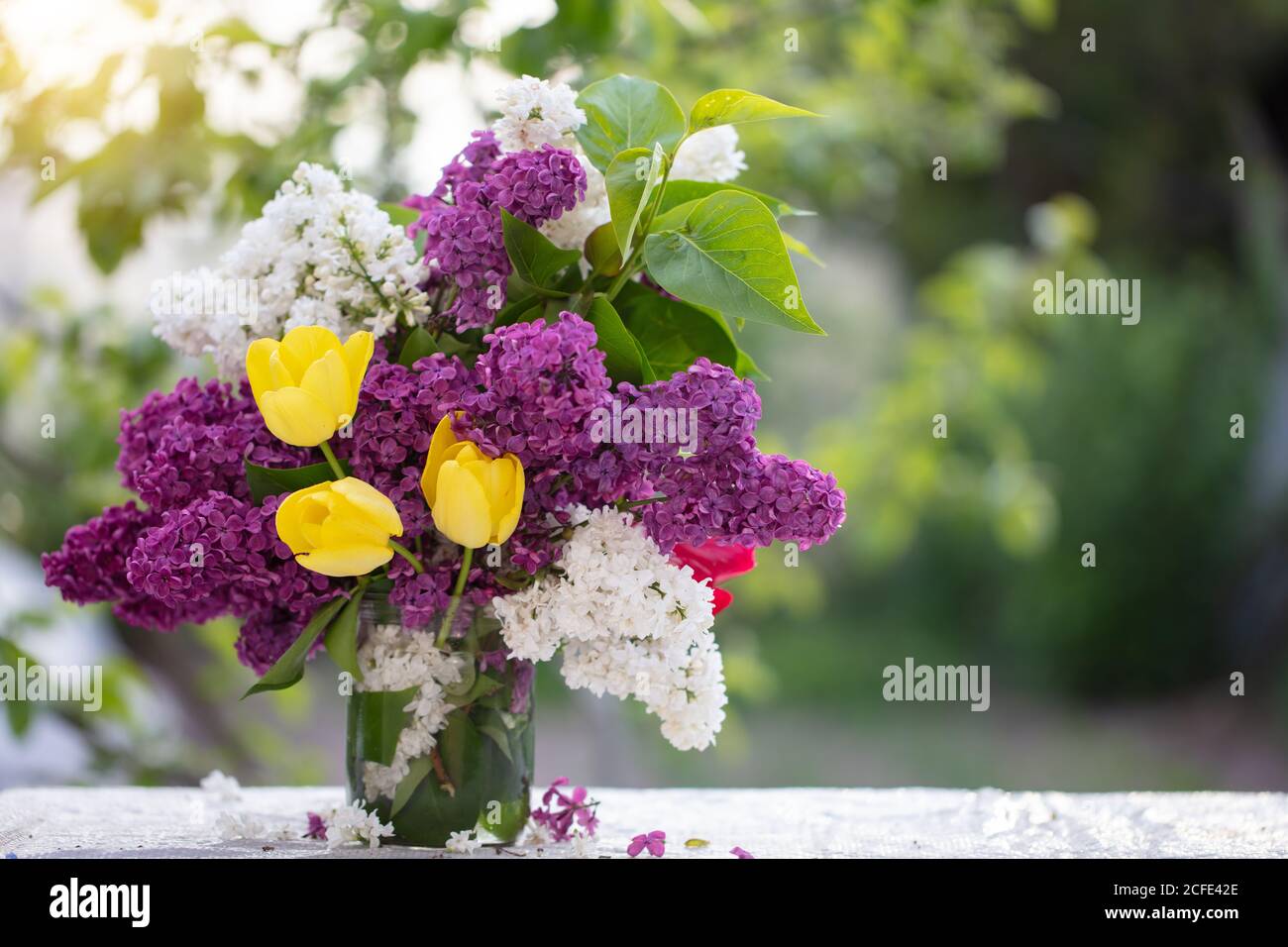 Printemps ou été. Un bouquet de lilas et de tulipes jaunes dans un pot en verre sur la table. Belles fleurs sur fond vert. Banque D'Images