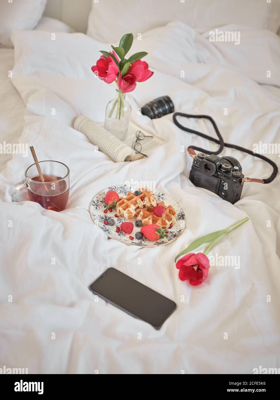 De dessus plat avec de délicieuses gaufres et baies fraîches sur le lit avec appareil photo, livre et téléphone Banque D'Images