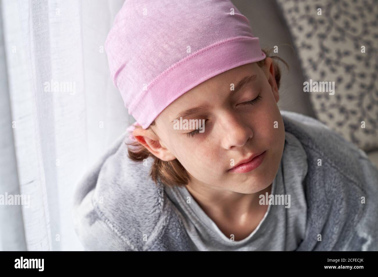 Enfant mignon sérieux dans le bandana rose avec les yeux fermés de combat cancer à la maison assis dans un canapé Banque D'Images
