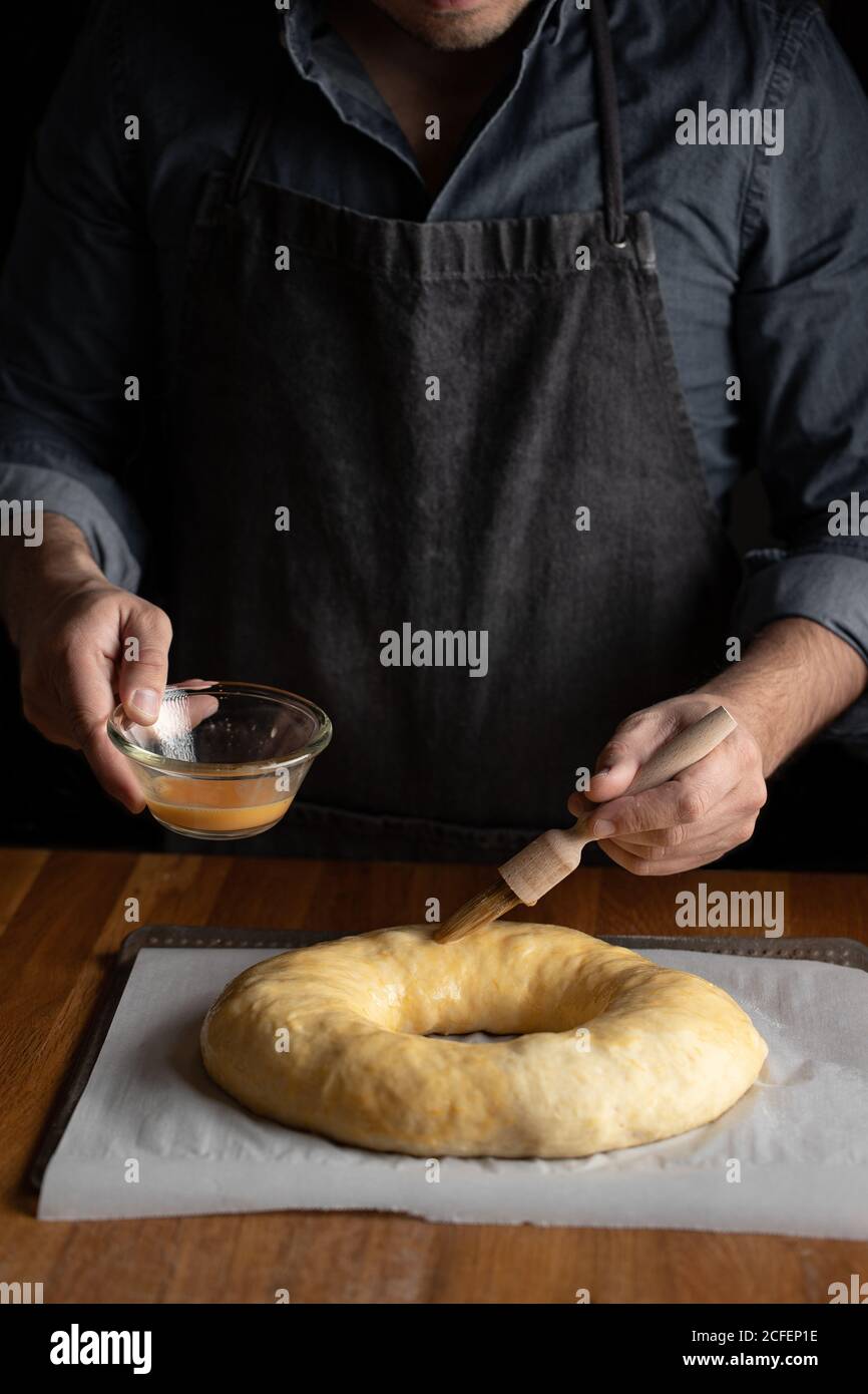 Petit chef cuisinier en tablier noir graissant du pain rond non cuit avec jaune d'œuf tout en étant debout sur une table en bois Banque D'Images