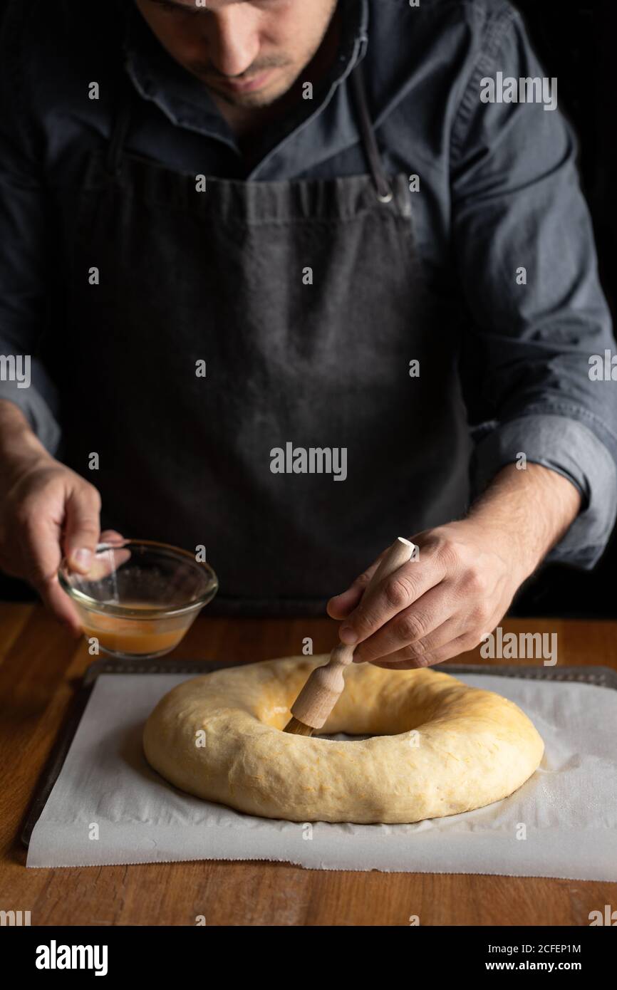 Petit chef cuisinier en tablier noir graissant du pain rond non cuit avec jaune d'œuf tout en étant debout sur une table en bois Banque D'Images