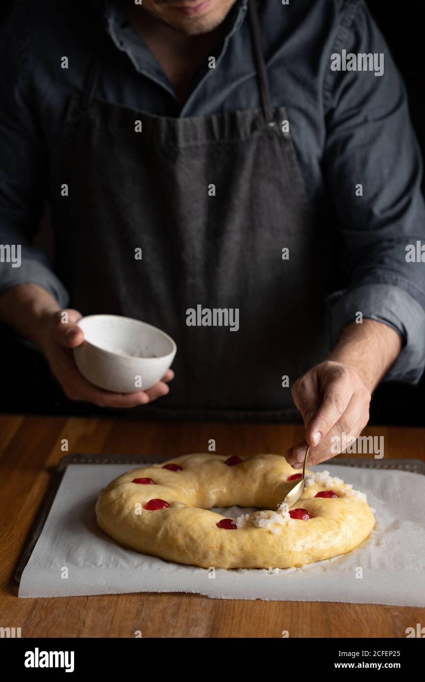 Chef cuisinier en tablier noir en graissant du pain rond non cuit avec jaune d'œuf recouvert de cerise tout en étant debout sur une table en bois Banque D'Images