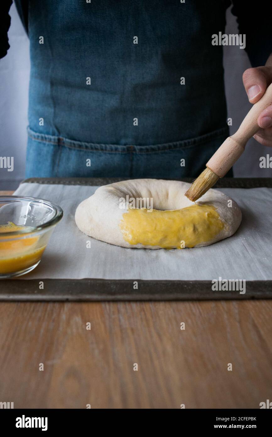 homme anonyme en tablier utilisant la brosse pour couvrir la pâte fraîche Avec jaune d'œuf tout en préparant le Rosca de Reyes dans la cuisine Banque D'Images