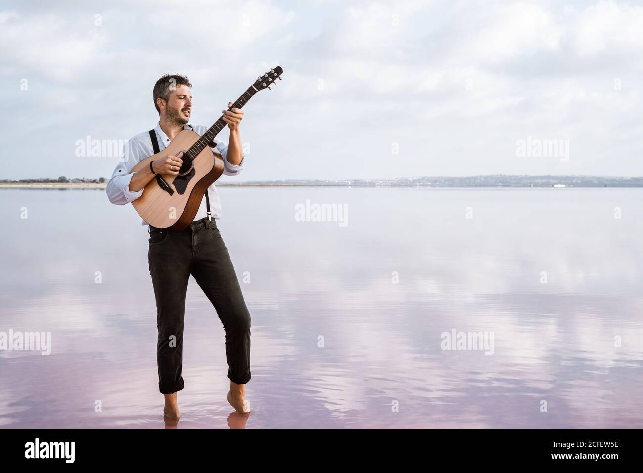Homme passionné en chemise blanche et bretelles jouant de la guitare  pendant se tenir pieds nus dans l'eau en bord de mer par temps nuageux  Photo Stock - Alamy