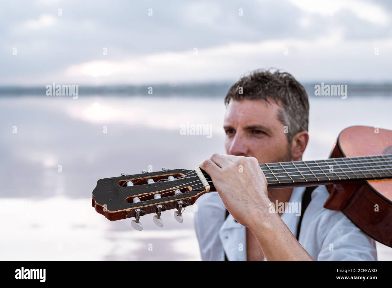 Homme plein de gite en chemise blanche et bretelles portant une guitare  acoustique et s'asseoir sur la plage avec l'eau rose vue loin entouré avec  une mer douce reflétant un paysage de