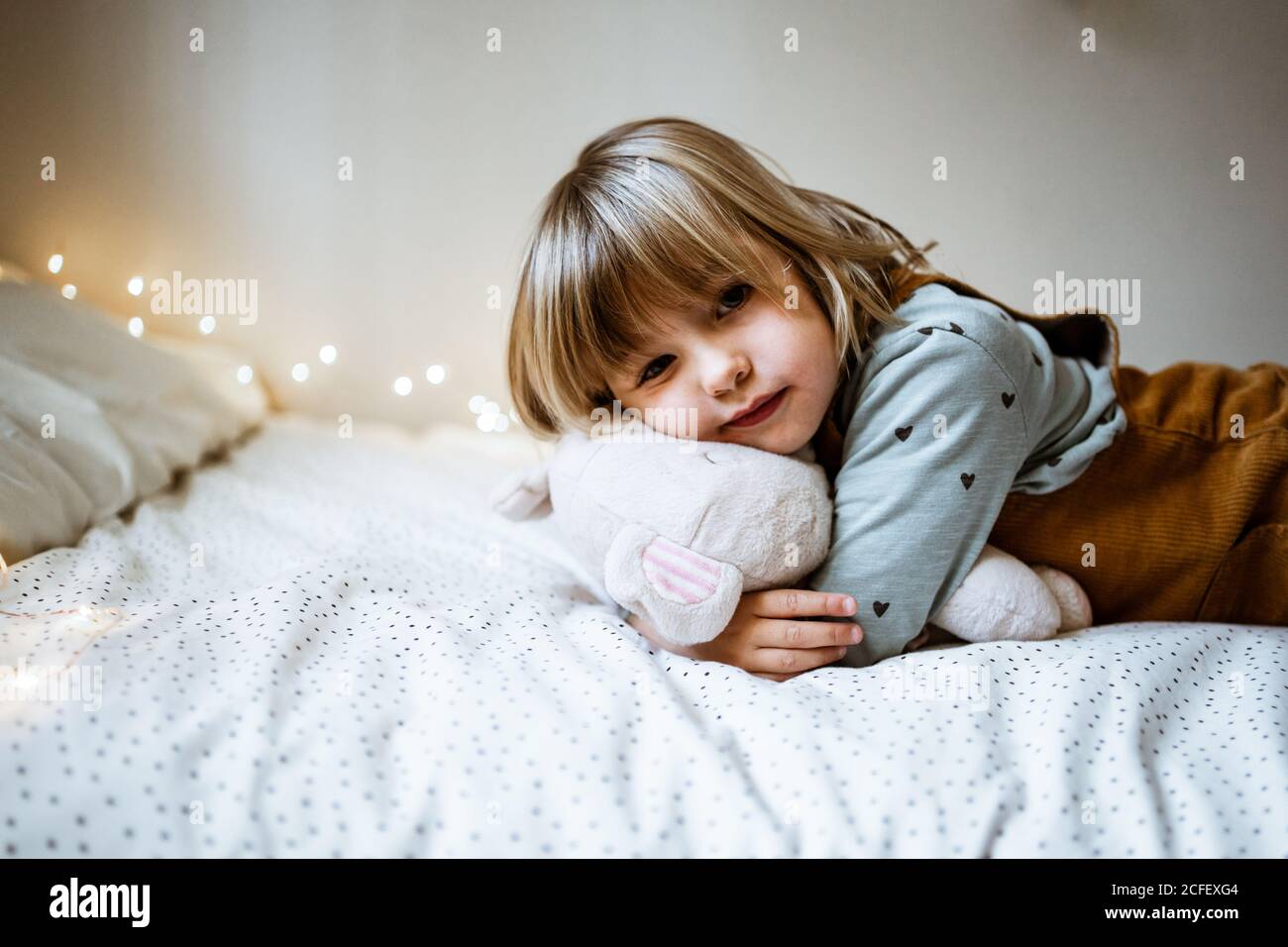 Une petite fille excitée embrasse un jouet en peluche et rit en mentant sur un lit mou près des lumières de fées à la maison Banque D'Images