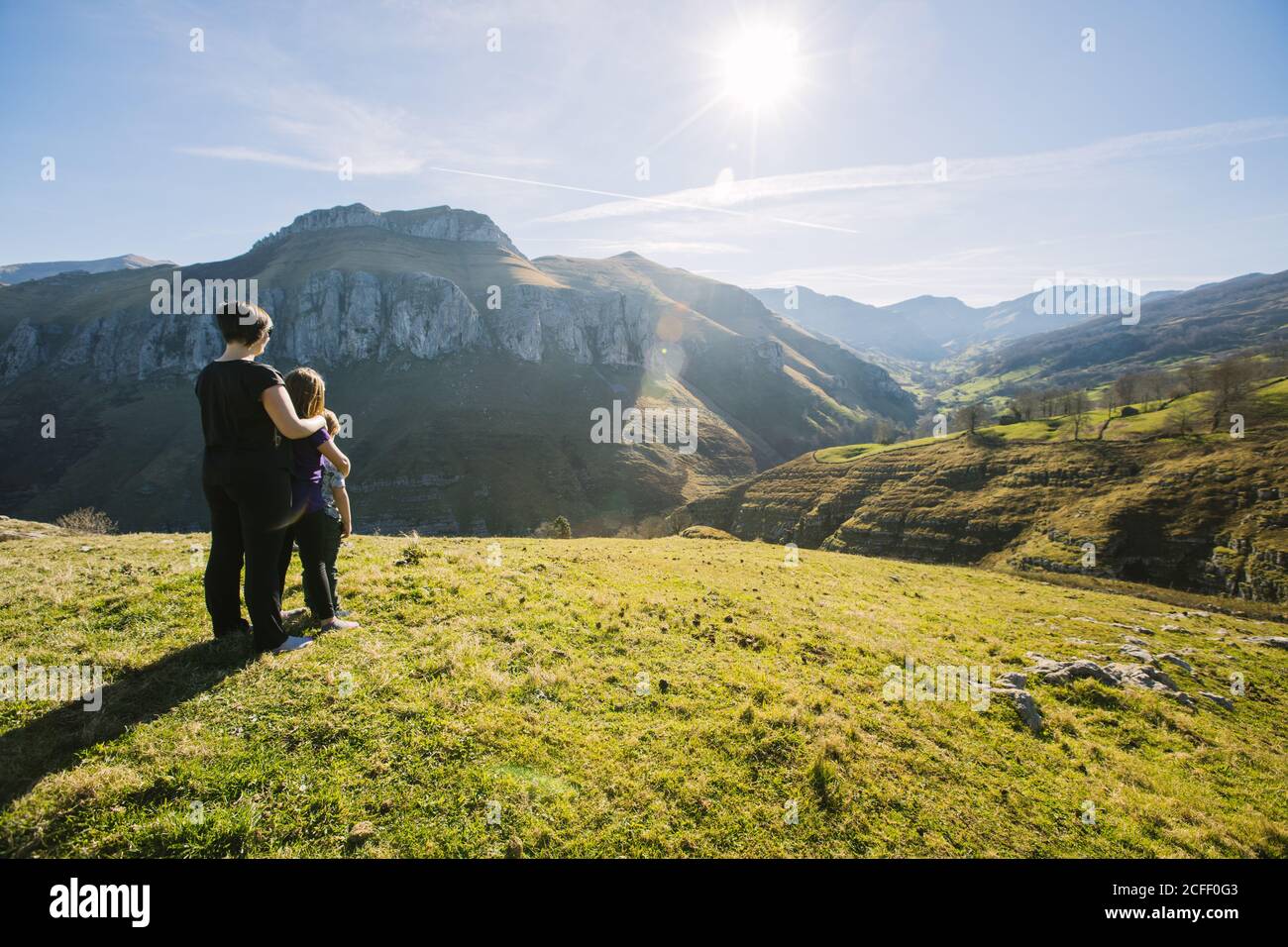 Vue arrière d'une randonneur anonyme qui embrasse les enfants en étant debout sur la prairie verte et l'observation pittoresque paysage de montagnes sur Journée ensoleillée en Cantabrie Banque D'Images