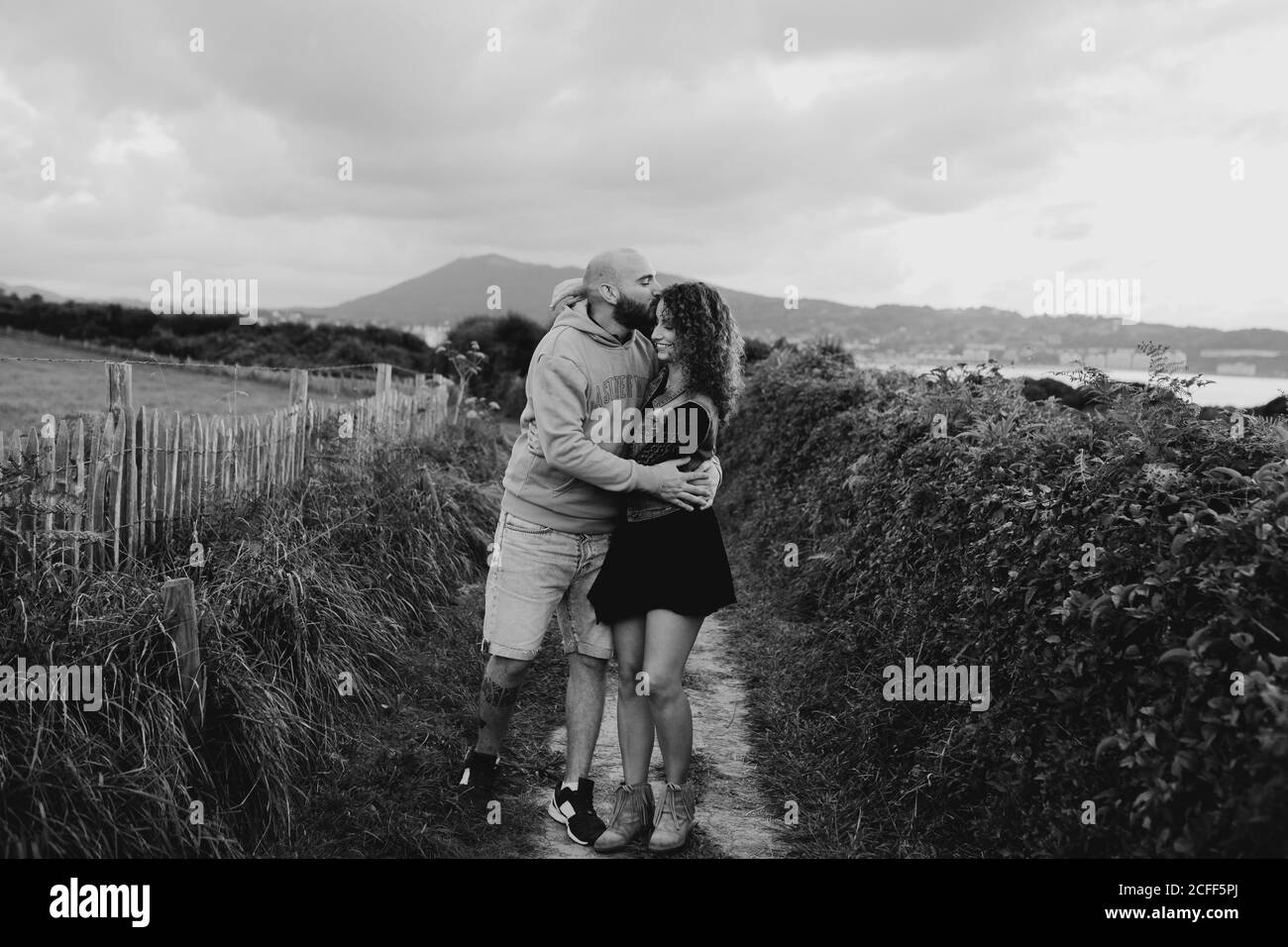 Femme tendre aimante touchant le menton de l'homme bien-aimé tout en marchant ensemble le long de la ferme verte en bord de mer avec des bâtiments de montagne et de ville en arrière-plan Banque D'Images