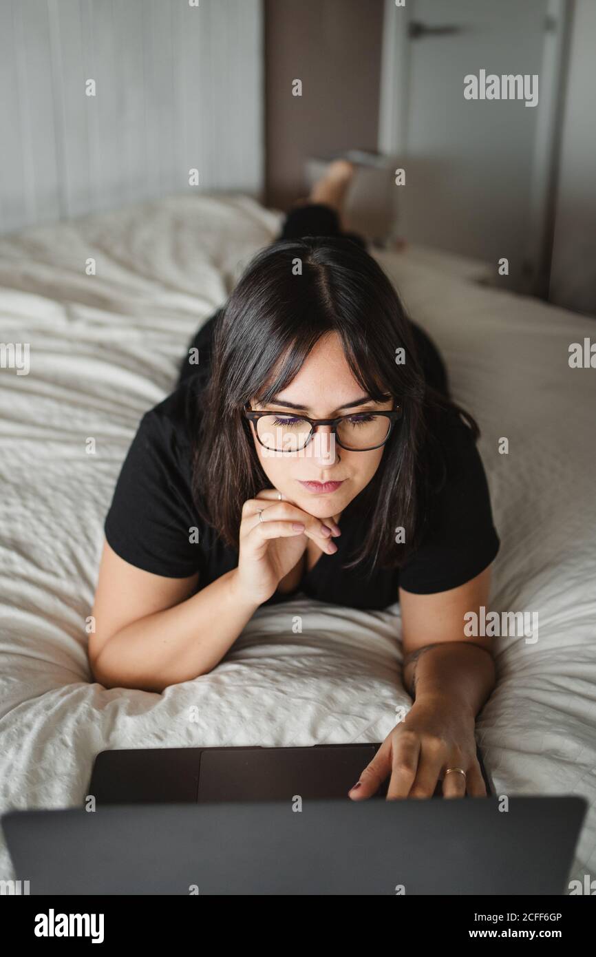 D'en haut de la vue de la jeune femme concentrée dans les lunettes dactylographiant sur ordinateur portable tout en étudiant et en posant sur le lit à la maison à Paris Banque D'Images