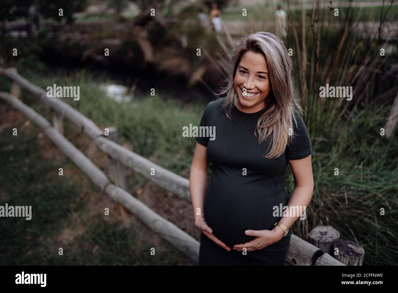 Belle femme enceinte en robe décontractée souriant et tenant les mains sur le ventre en se tenant à côté d'un pont en bois sur le petit rivière dans le parc en été en regardant la caméra Banque D'Images