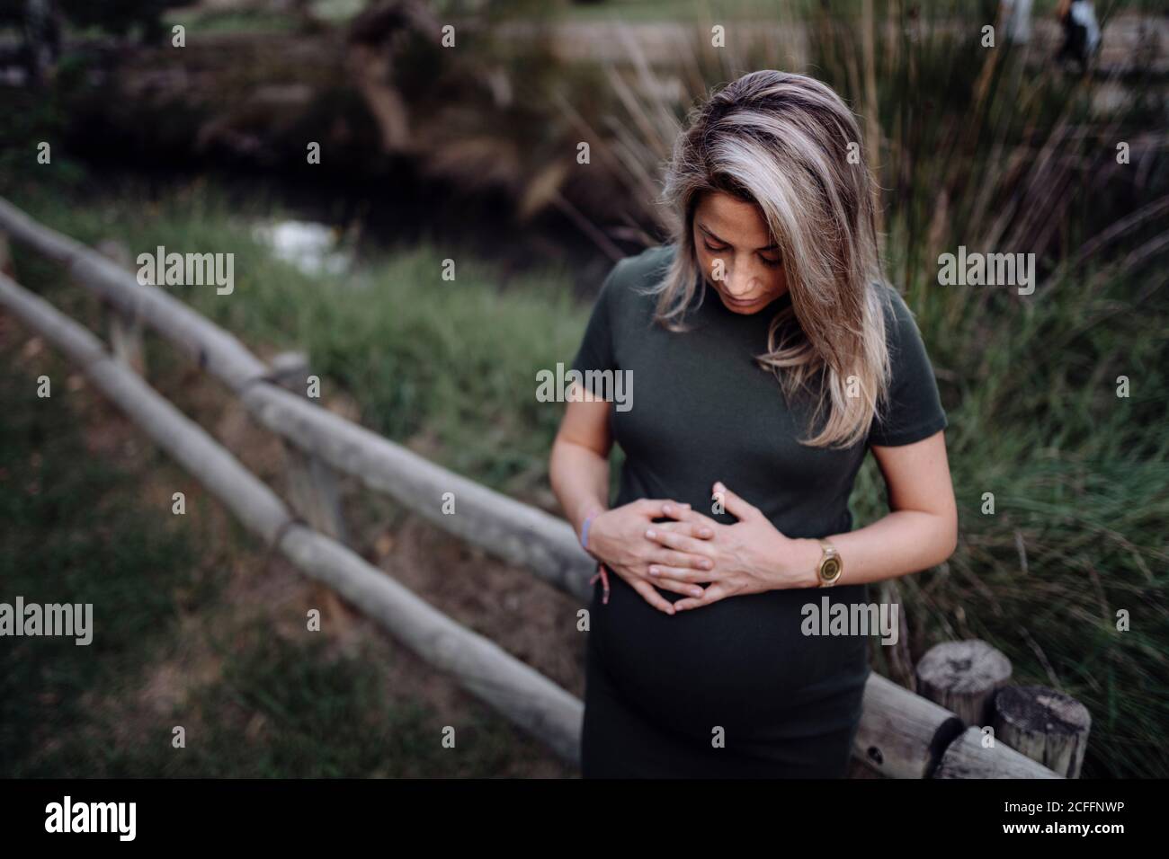 Belle femme enceinte en robe décontractée souriant et tenant les mains sur le ventre en se tenant à côté d'un pont en bois sur le petit rivière dans le parc en été Banque D'Images