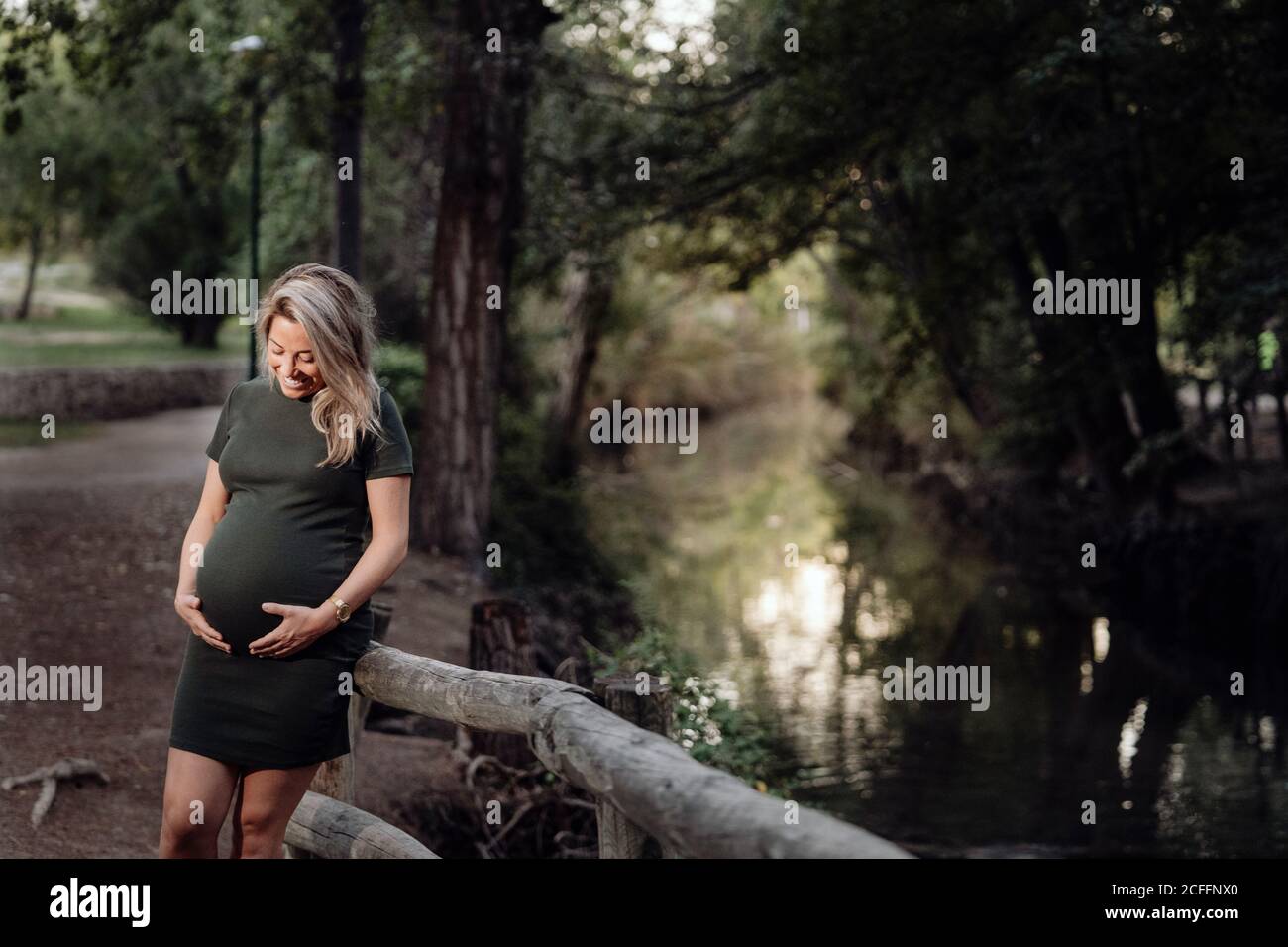 Belle femme enceinte en robe décontractée souriant et tenant les mains sur le ventre en se tenant à côté d'un pont en bois sur le petit rivière dans le parc en été Banque D'Images