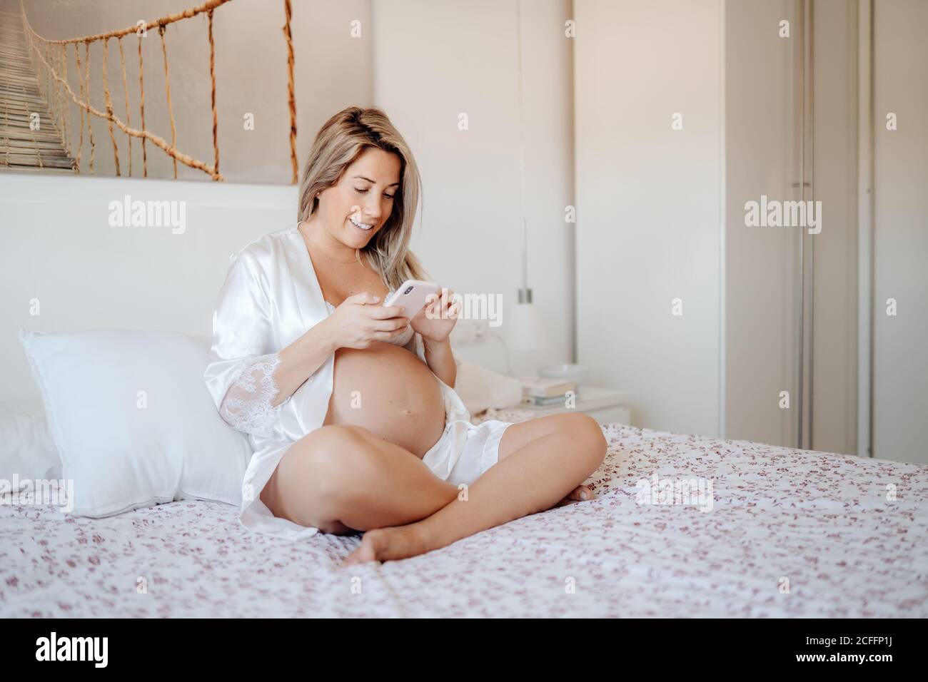 Contenu blonde femme enceinte en blanc ouvert chemise et soutien-gorge navigation sur votre téléphone mobile en vous asseyant avec des jambes croisées sur de grandes lit dans une chambre lumineuse Banque D'Images