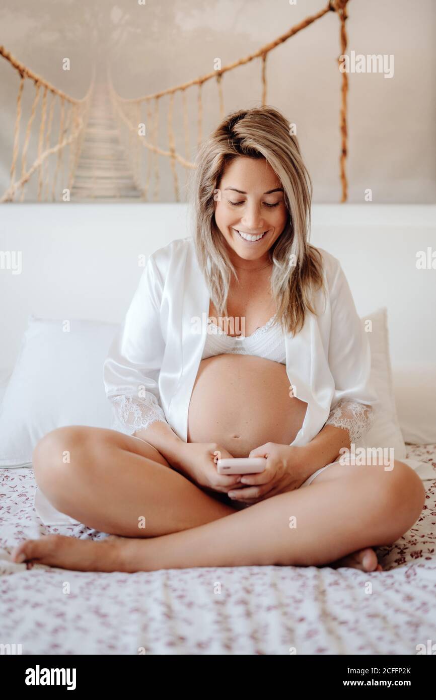 Contenu blonde femme enceinte en blanc ouvert chemise et soutien-gorge navigation sur votre téléphone mobile en vous asseyant avec des jambes croisées sur de grandes lit dans une chambre lumineuse Banque D'Images