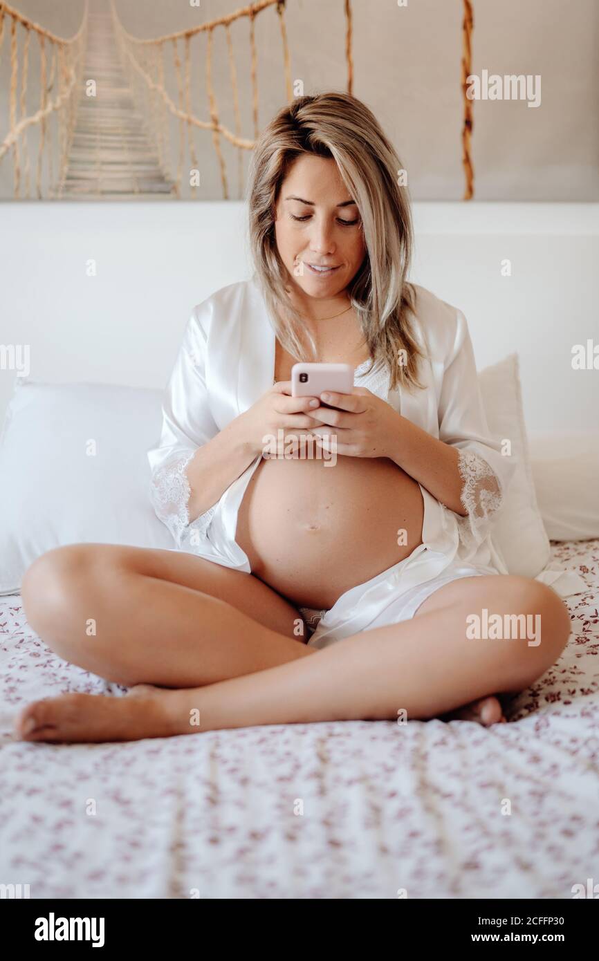 Contenu blonde femme enceinte en blanc ouvert chemise et soutien-gorge navigation sur votre téléphone mobile en vous asseyant avec des jambes croisées sur de grandes lit dans une chambre lumineuse Banque D'Images