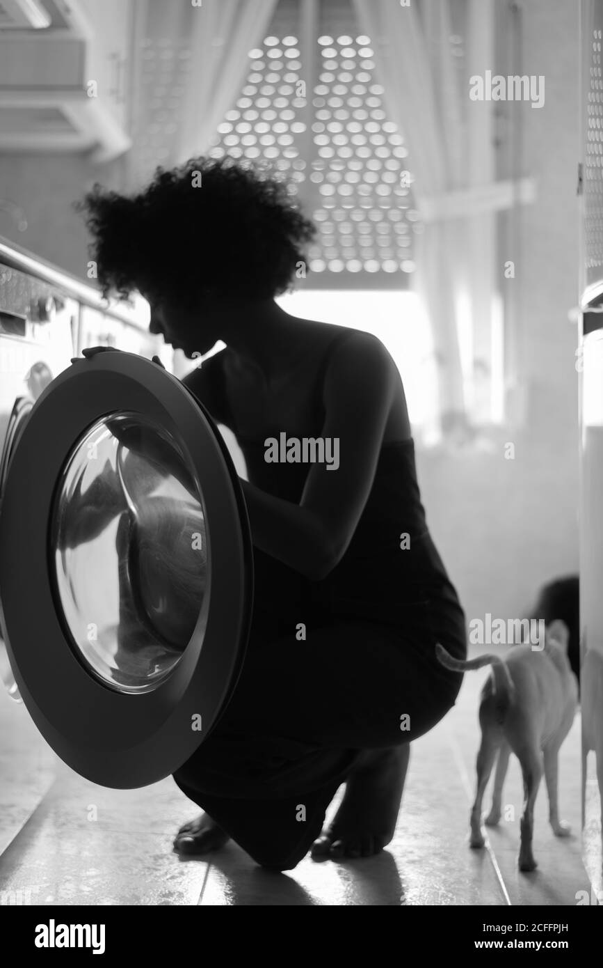 Vue latérale de la femme de ménage ethnique noir et blanc en train de charger le linge machine avec des vêtements sales à la maison Banque D'Images