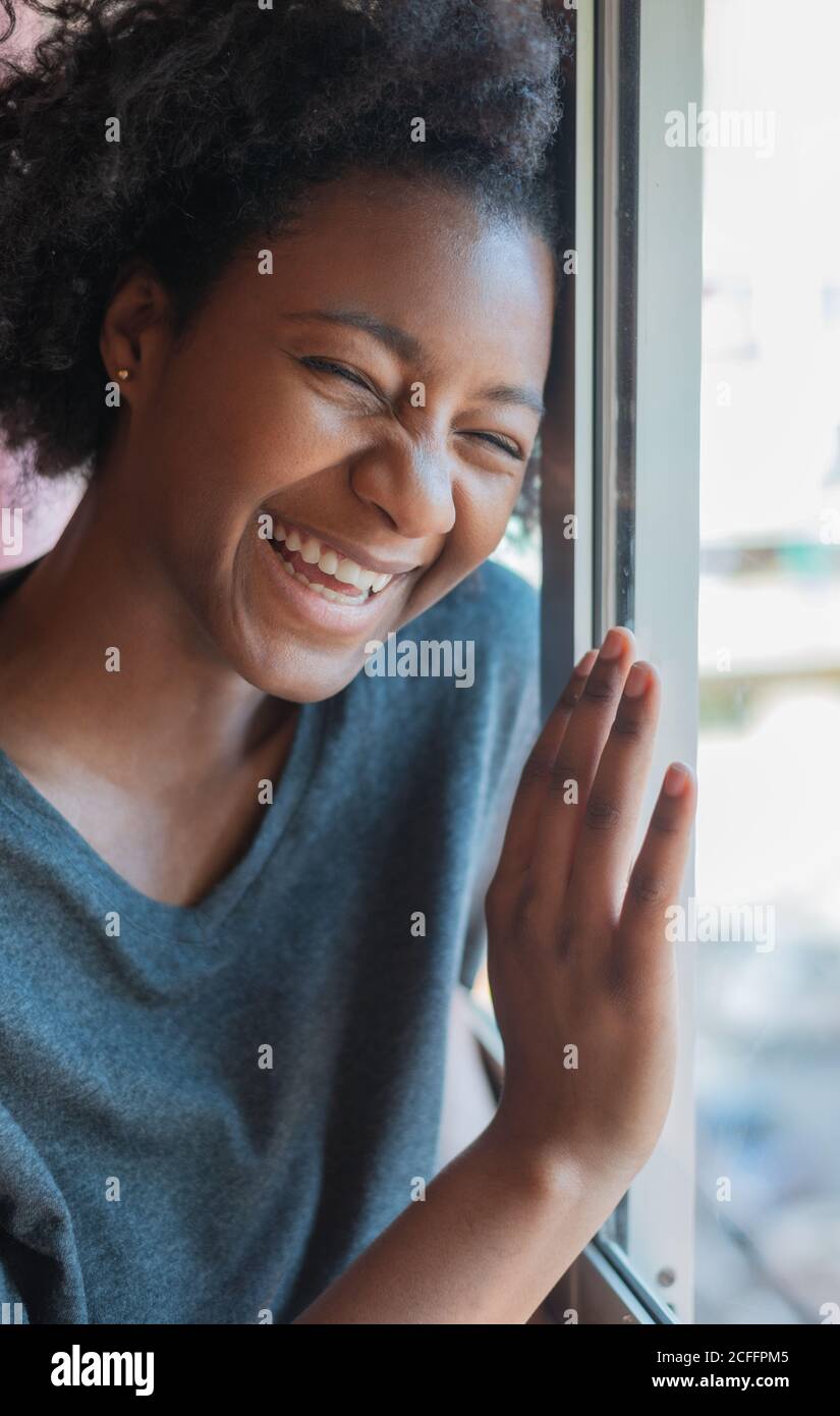 Gaie ethnique femme dans une tenue décontractée penchée sur la fenêtre et riant à la blague avec les yeux fermés tout en se reposant pendant le week-end à la maison Banque D'Images