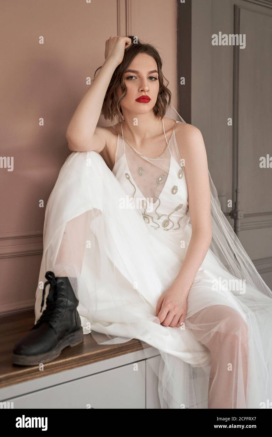 Femme provocatrice aux lèvres rouges dans la robe de mariage blanche et des  chaussures noires brutales assis sur la poitrine des tiroirs penchée sur la  main et regardant l'appareil photo contre le