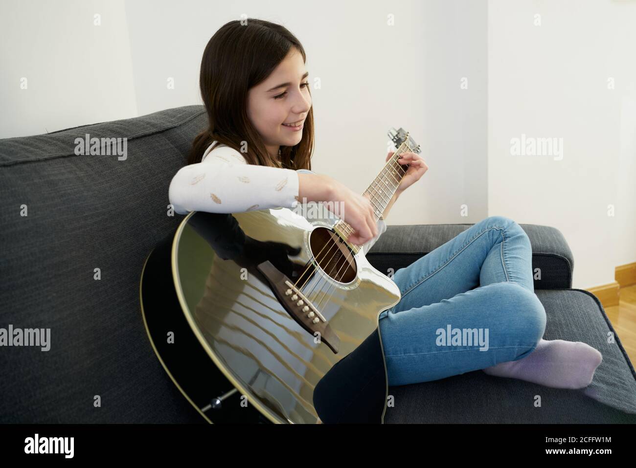 Jeune fille gaie dans des vêtements décontractés assis sur un canapé confortable avec les jambes croisées et jouer de la guitare acoustique tout en passant du temps à la maison Banque D'Images