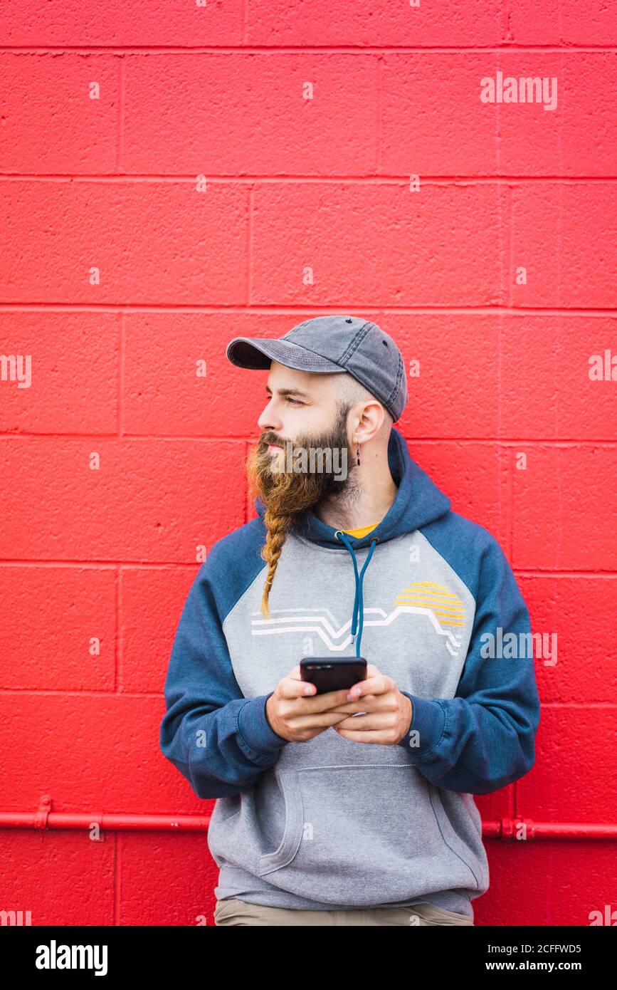 Un homme séduisant avec une barbe tressée qui navigue sur un smartphone  tout en se penchant rouge mur sur la rue de la ville Photo Stock - Alamy