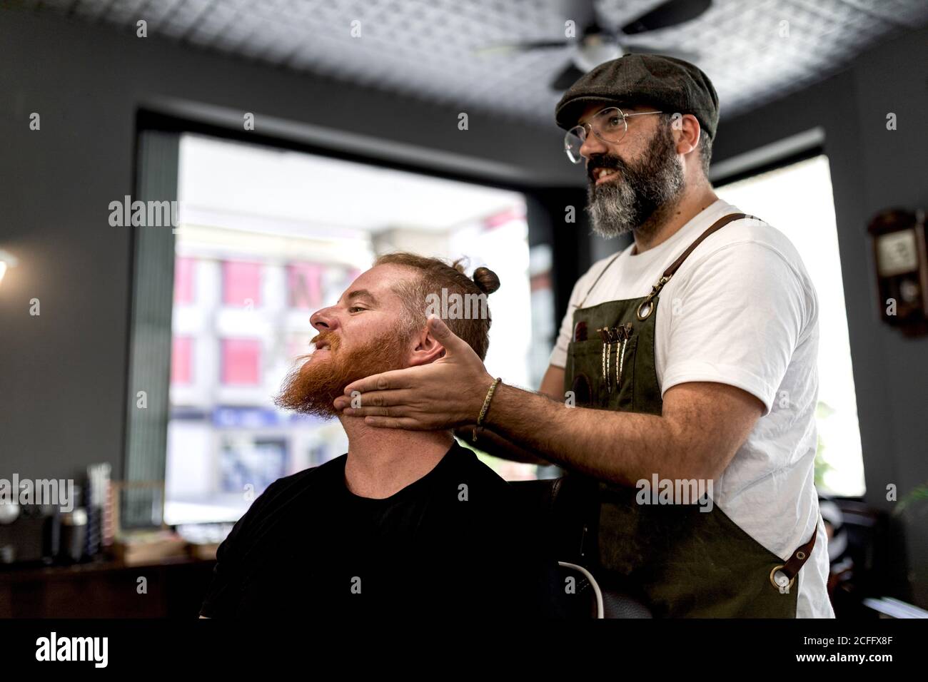 Vue latérale du coiffeur faisant un massage du visage à la belle tête rouge homme assis sur une chaise Banque D'Images