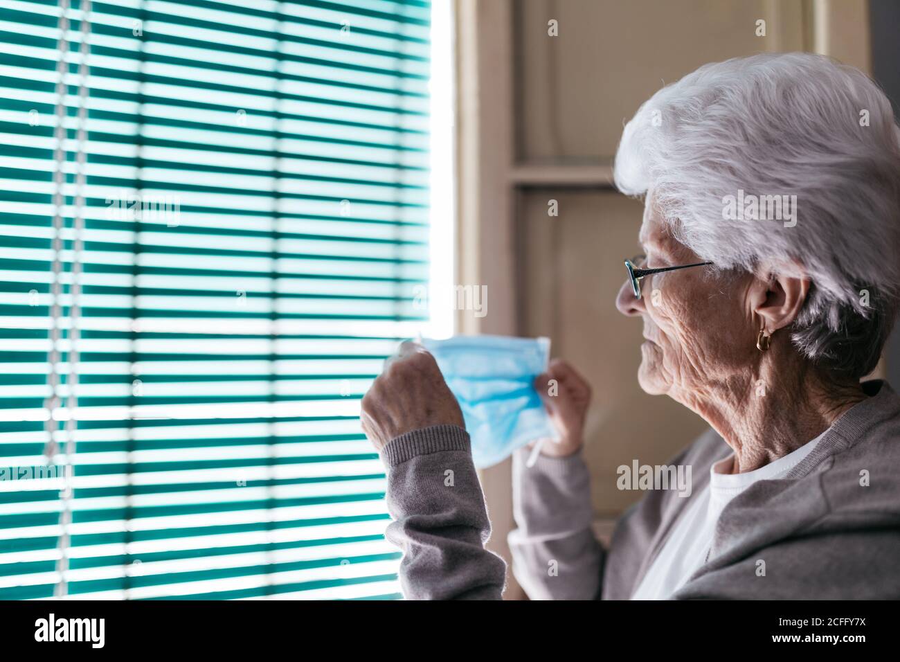 Vue latérale de la femme à cheveux gris se préparant à porter un appareil médical masque pour la prévention du coronavirus en se tenant près de la fenêtre avec obturateur à accueil Banque D'Images