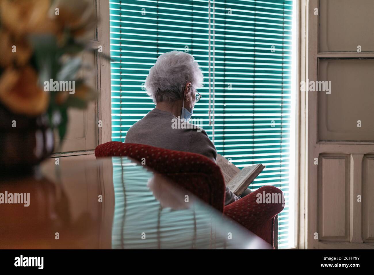 Femme senior dans le masque de protection et les gants de lecture livre à accueil Banque D'Images