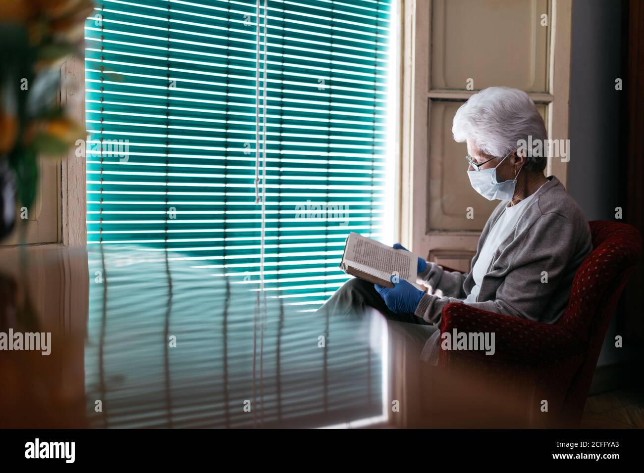 Femme senior dans le masque de protection et les gants de lecture livre à accueil Banque D'Images
