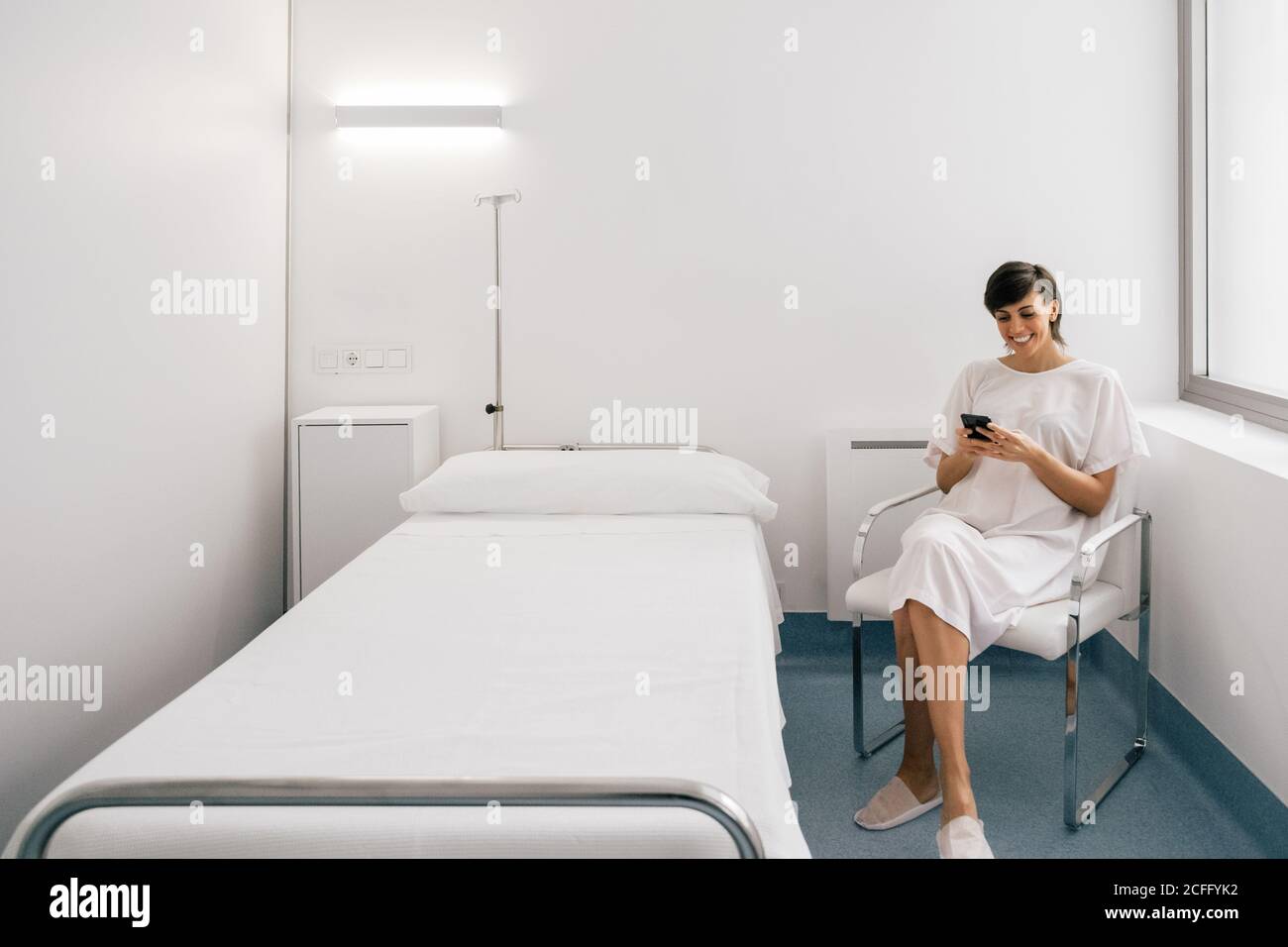 Femme gaie en robe blanche assise sur le lit près du lit et de la navigation de smartphone dans la salle de clinique moderne Banque D'Images